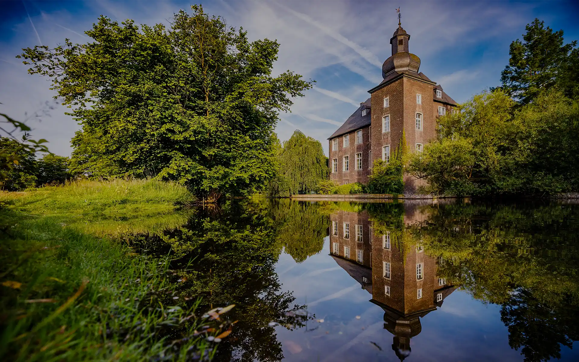 Historisches Backstein-Wasserschloss, umgeben von Bäumen, spiegelt sich klar in einem ruhigen Teich unter blauem Himmel.